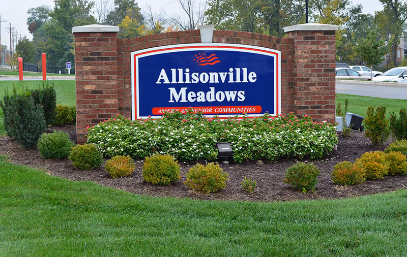 Brick entrance sign that reads 'Allisonville Meadows' set in a landscaped bed of shrubs and flowers.