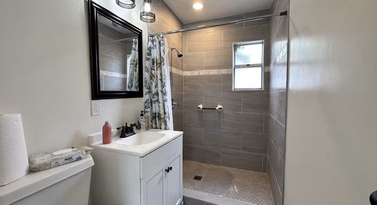 A clean bathroom featuring a white vanity with a sink, a mirror above it, a roll of paper towels and a pack of wipes on the toilet tank, and a walk-in shower with gray tiles, a floral shower curtain, a small window, and a grab bar.