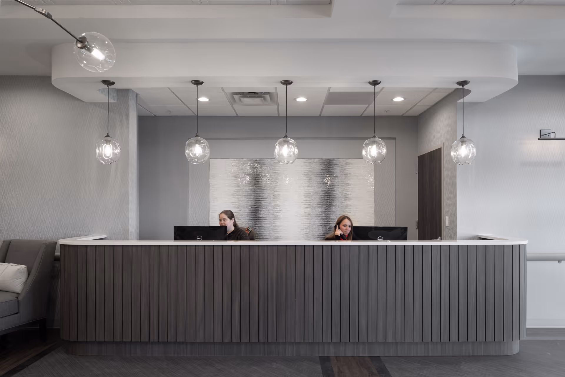 Reception area with a modern wooden front desk and two receptionists working behind computers. The background features a textured silver wall art and five hanging glass pendant lights. A gray sofa with a white pillow is partially visible on the left side.