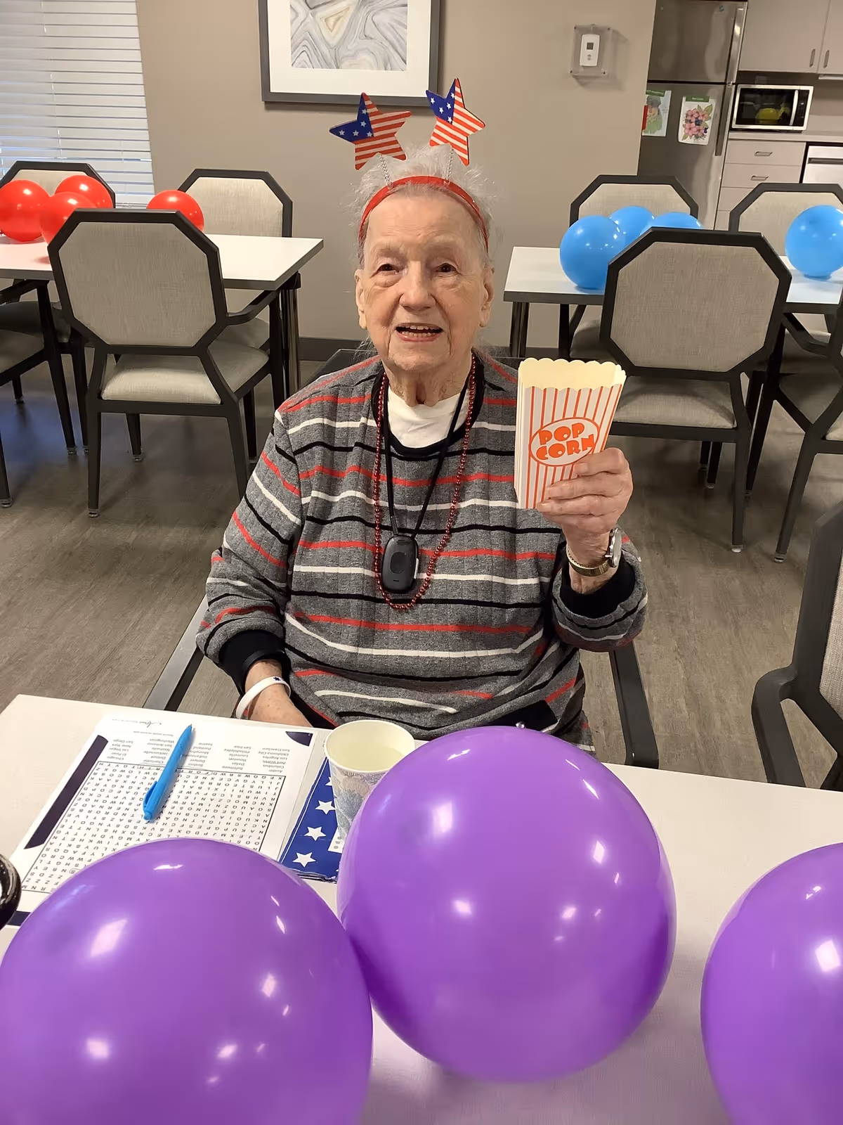 An elderly woman wearing a striped sweater and a headband with red, white, and blue star decorations is sitting at a table in a dining area. She is holding a small popcorn container and smiling. The table has purple balloons, a paper cup, and a word search puzzle with a blue pen. In the background, there are chairs, tables with red and blue balloons, and a kitchen area.