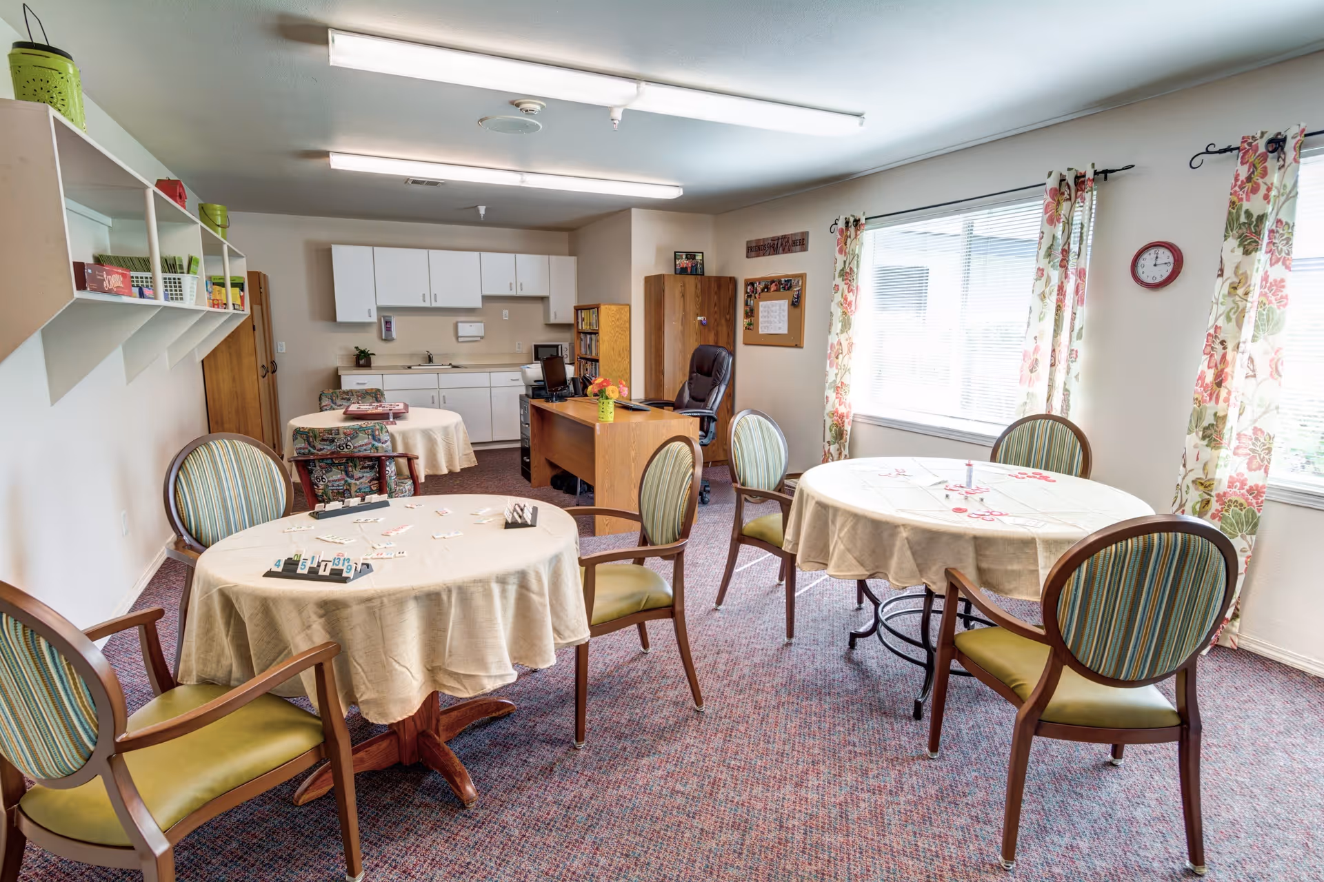 A bright room with round tables covered in beige tablecloths and surrounded by wooden chairs with green and striped cushions. The room has floral curtains on the windows, a desk with a computer and office chair, white cabinets along the back wall, and shelves with various items. The carpet is patterned and the room is well-lit with fluorescent ceiling lights.
