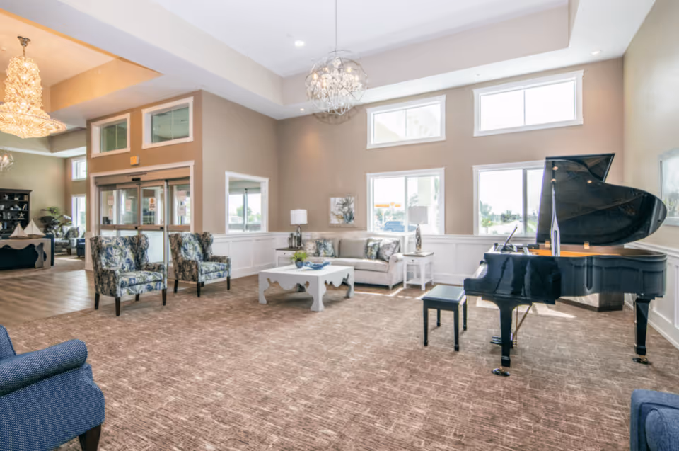 A spacious and well-lit living room area in a senior living facility featuring a black grand piano with a matching bench, a beige sofa with patterned cushions, two floral armchairs, a white coffee table, and large windows allowing natural light to fill the room. The room has beige walls, carpeted flooring, and modern chandeliers hanging from the ceiling.