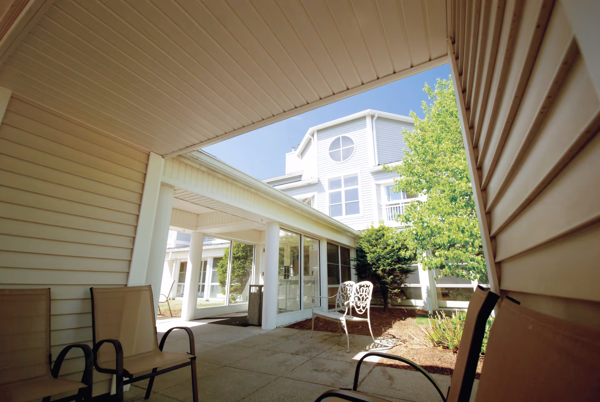 Covered outdoor walkway and seating area leading to a white multi-story senior living building with windows and landscaping.