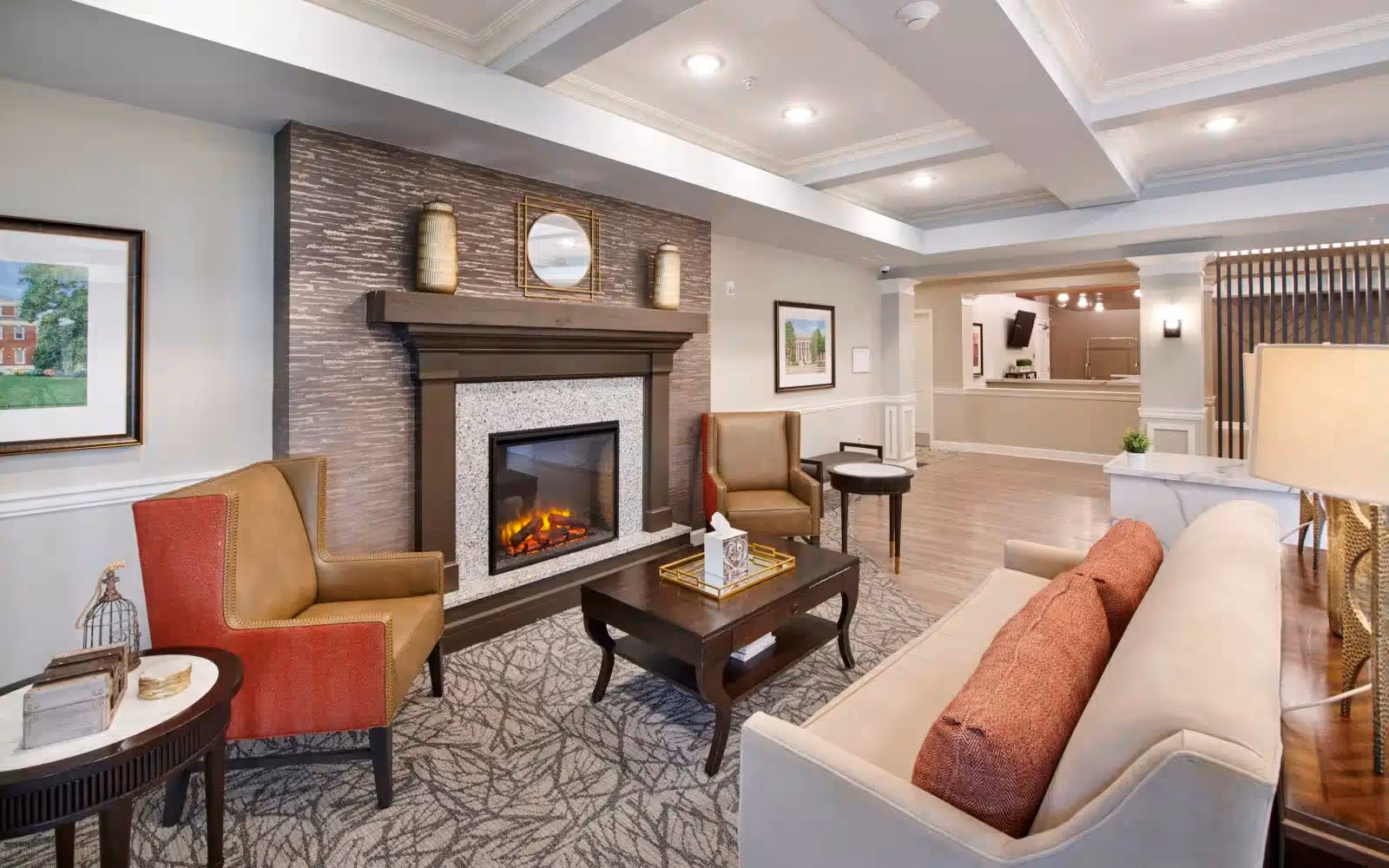A cozy living room area in a senior living facility featuring a beige sofa with orange cushions, two armchairs with red and tan upholstery, a dark wooden coffee table with a tissue box on a tray, and a fireplace with a decorative mantle and a round mirror above it. The room has patterned carpet, framed artwork on the walls, and recessed ceiling lights.