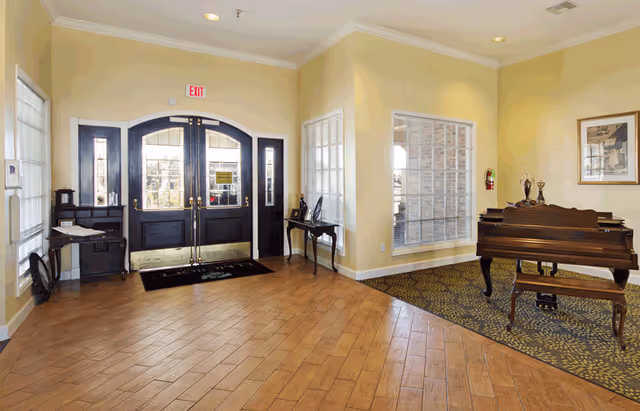 Entrance area of a senior living facility with double black doors featuring glass panels, a wooden floor, a small piano with a bench on a patterned carpet, a side table with decorative items, and large windows allowing natural light.