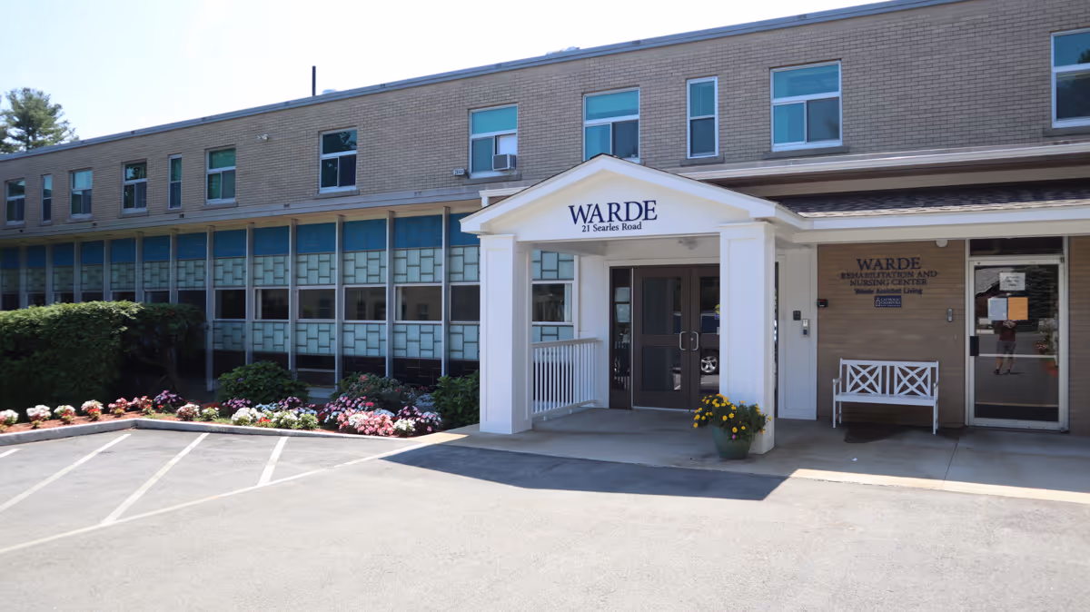 Exterior view of Warde Supported Residential Care building showing the entrance with a white portico labeled 'WARDE 21 Searles Road'. There are flower beds with colorful flowers along the front and a parking area in the foreground. The building is two stories with multiple windows and a bench near the entrance.