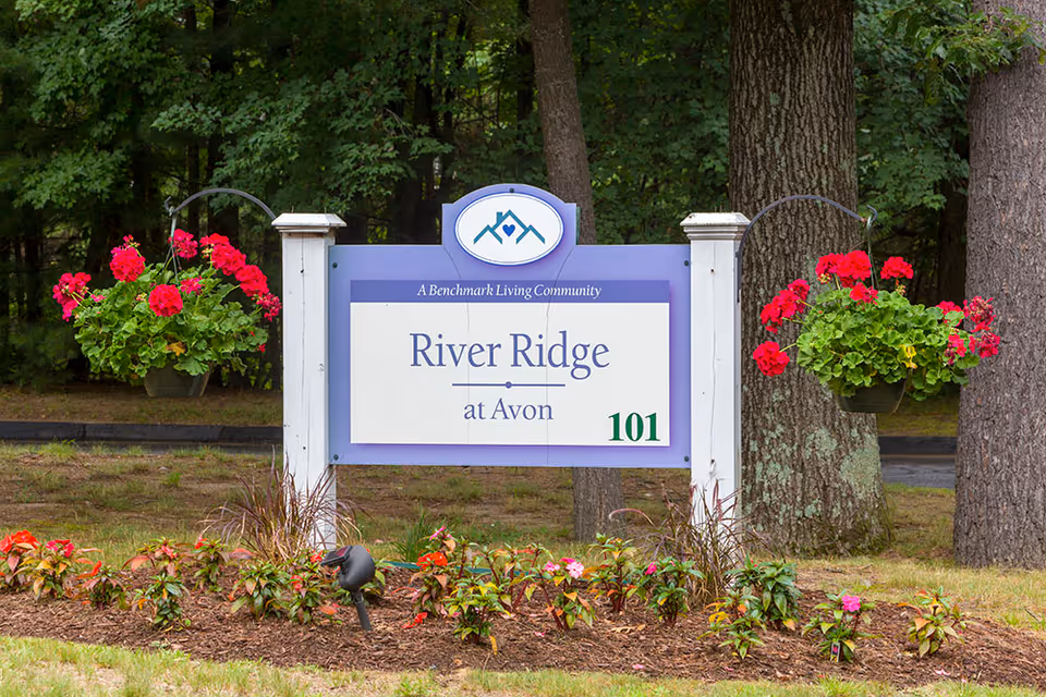 Outdoor entrance sign reading "River Ridge at Avon" with hanging flower baskets and landscaped beds in front of trees.