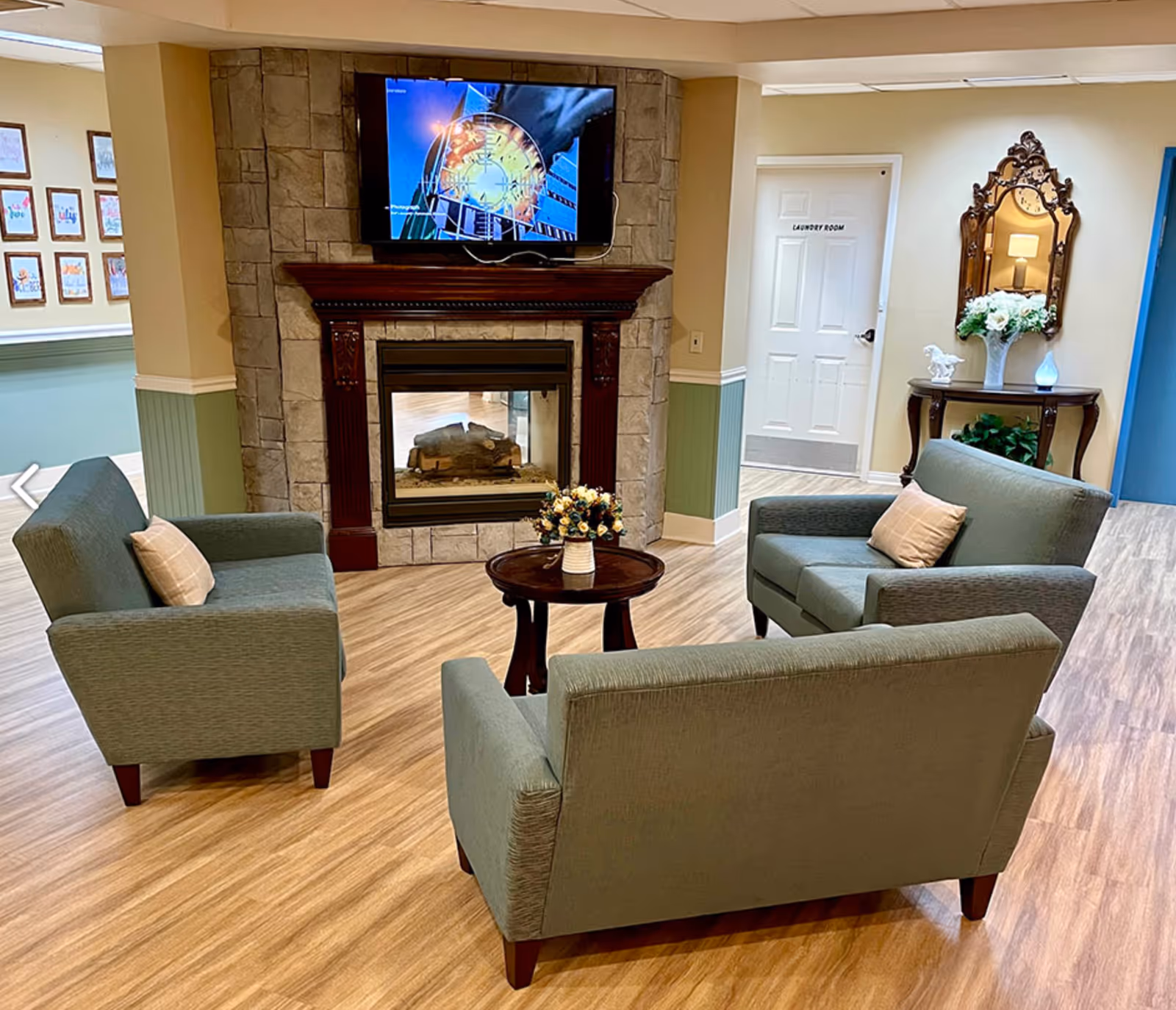 A cozy sitting area in a senior living facility with three green upholstered armchairs arranged around a small round wooden table with a flower vase. Behind the seating area is a stone fireplace with a mounted flat-screen TV above it. To the right, there is a decorative wooden console table with a mirror, flowers, and a lamp. A door labeled 'Laundry Room' is visible in the background.