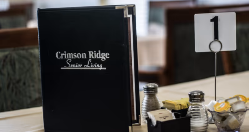 A close-up view of a dining table at Crimson Ridge Senior Living, featuring a black menu holder with the text 'Crimson Ridge Senior Living,' a table number holder displaying the number 1, salt and pepper shakers, and containers holding sugar packets and creamers.