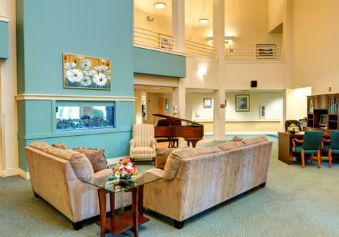 Community lounge with two beige sofas around a glass side table, a grand piano, a built-in aquarium, and a two-story open atrium.