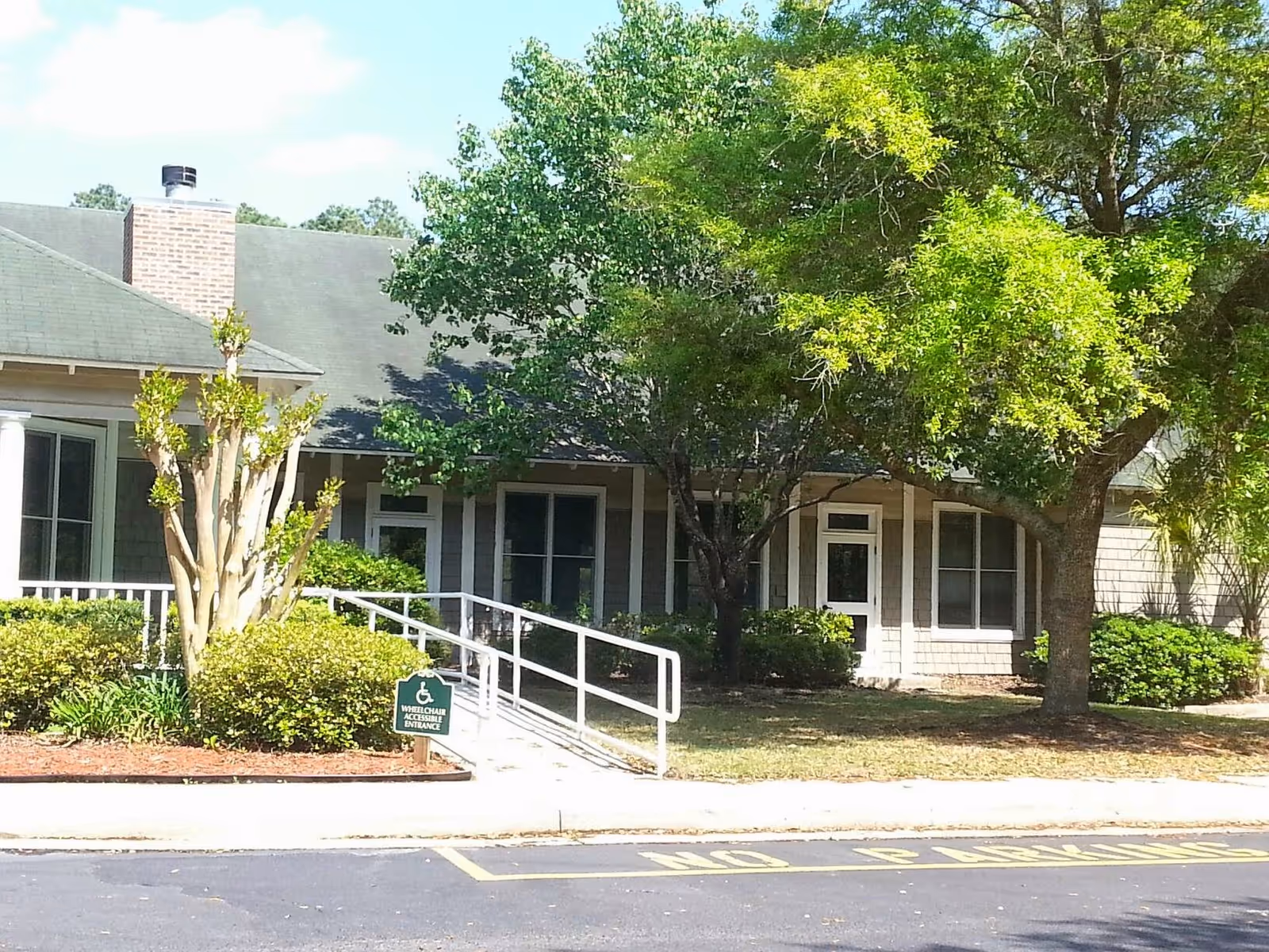Exterior view of a single-story building with a wheelchair accessible ramp leading to the entrance. The building is surrounded by green trees and bushes, with a sign indicating a wheelchair accessible entrance. The parking area in front has a 'No Parking' zone marked on the pavement.