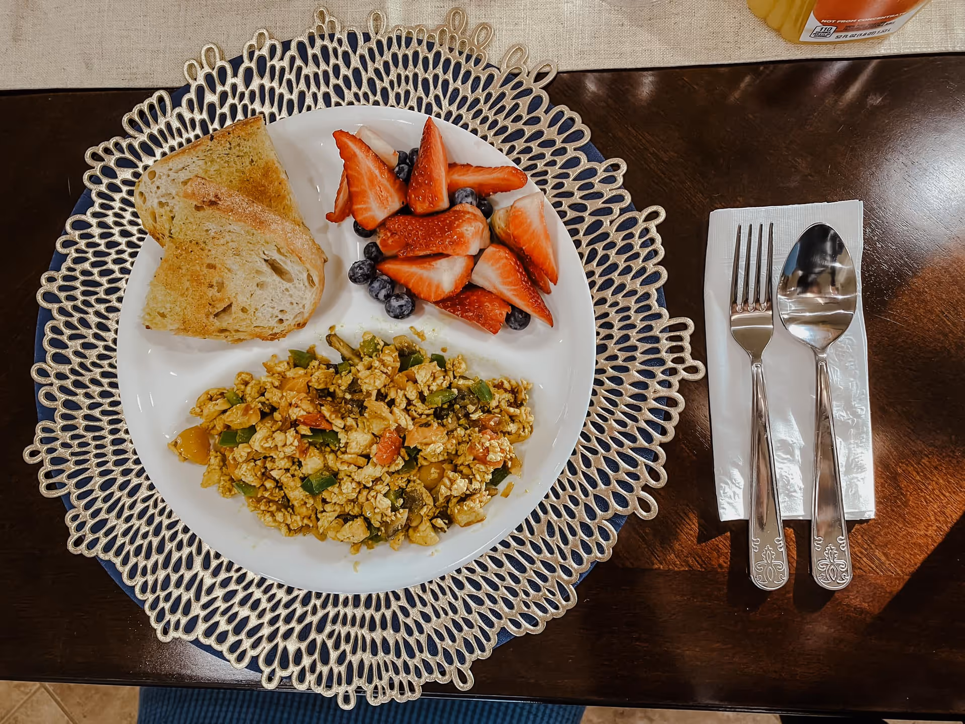 A white plate on a decorative placemat with scrambled eggs mixed with vegetables, two slices of toasted bread, and a serving of fresh strawberries and blueberries. To the right of the plate, there is a fork and spoon placed on a folded white napkin on a dark wooden table.