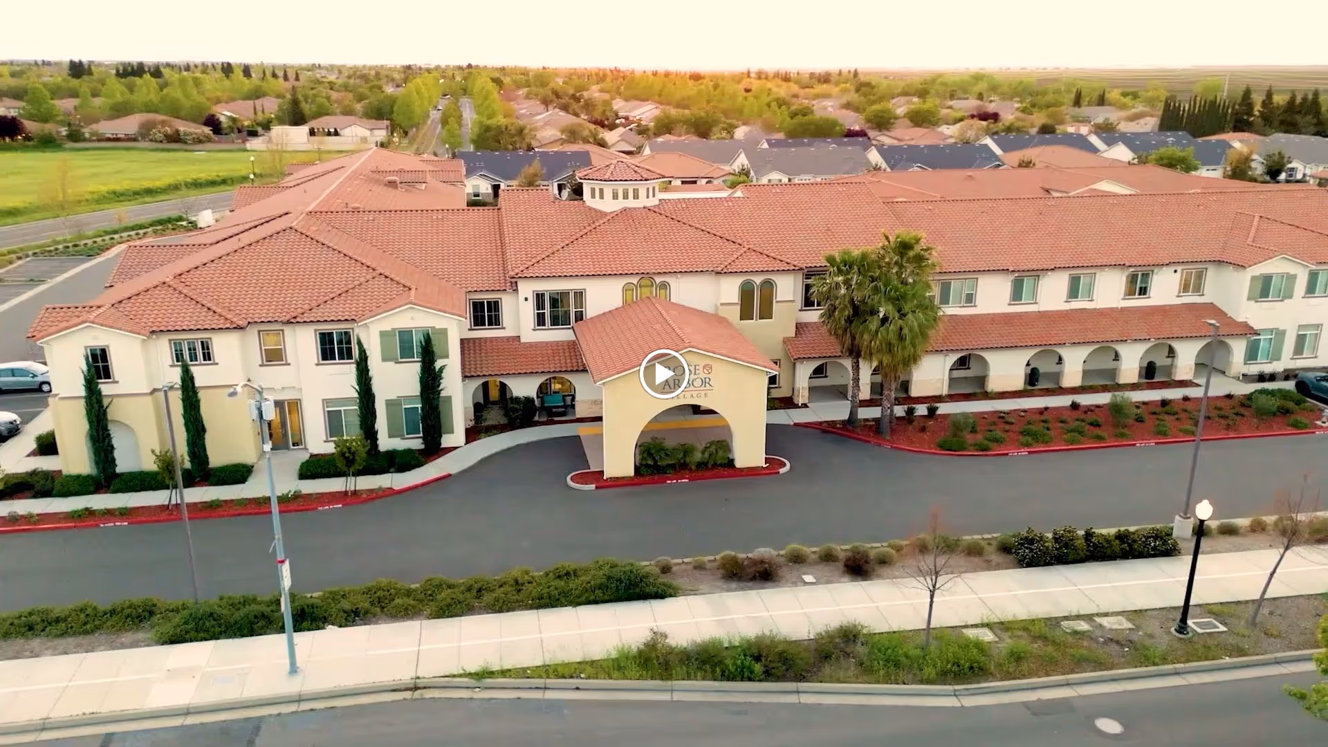 Aerial view of a large senior living facility building with a red-tiled roof, beige walls, and an arched entrance canopy. The building is surrounded by landscaped greenery, a driveway, and a sidewalk with street lamps. Residential houses and trees are visible in the background.