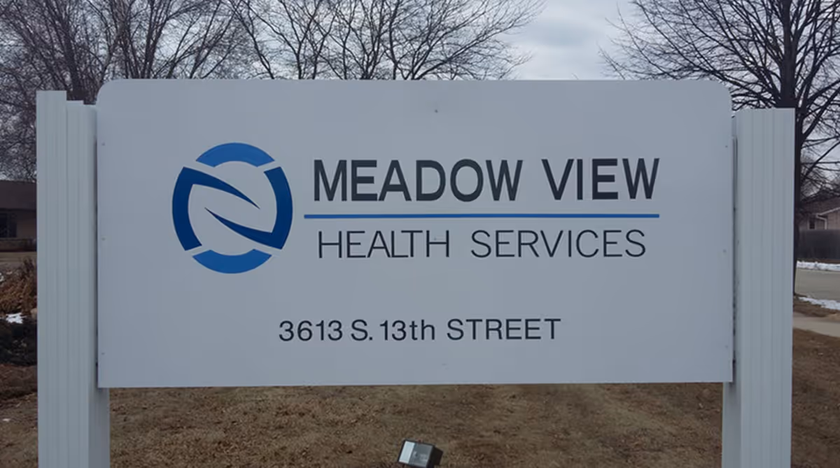 Outdoor sign for Meadow View Health Services located at 3613 S. 13th Street, with a blue and white logo on the left side. The sign is mounted on white posts with a background of leafless trees and a cloudy sky.