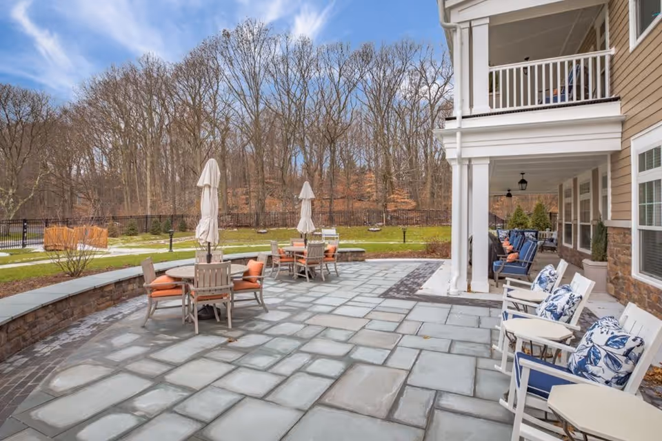 Outdoor patio area at Sunrise of Fairfield with stone tile flooring, several round tables with chairs and umbrellas, and a covered porch with cushioned seating. Bare trees and a grassy area are visible in the background under a partly cloudy sky.