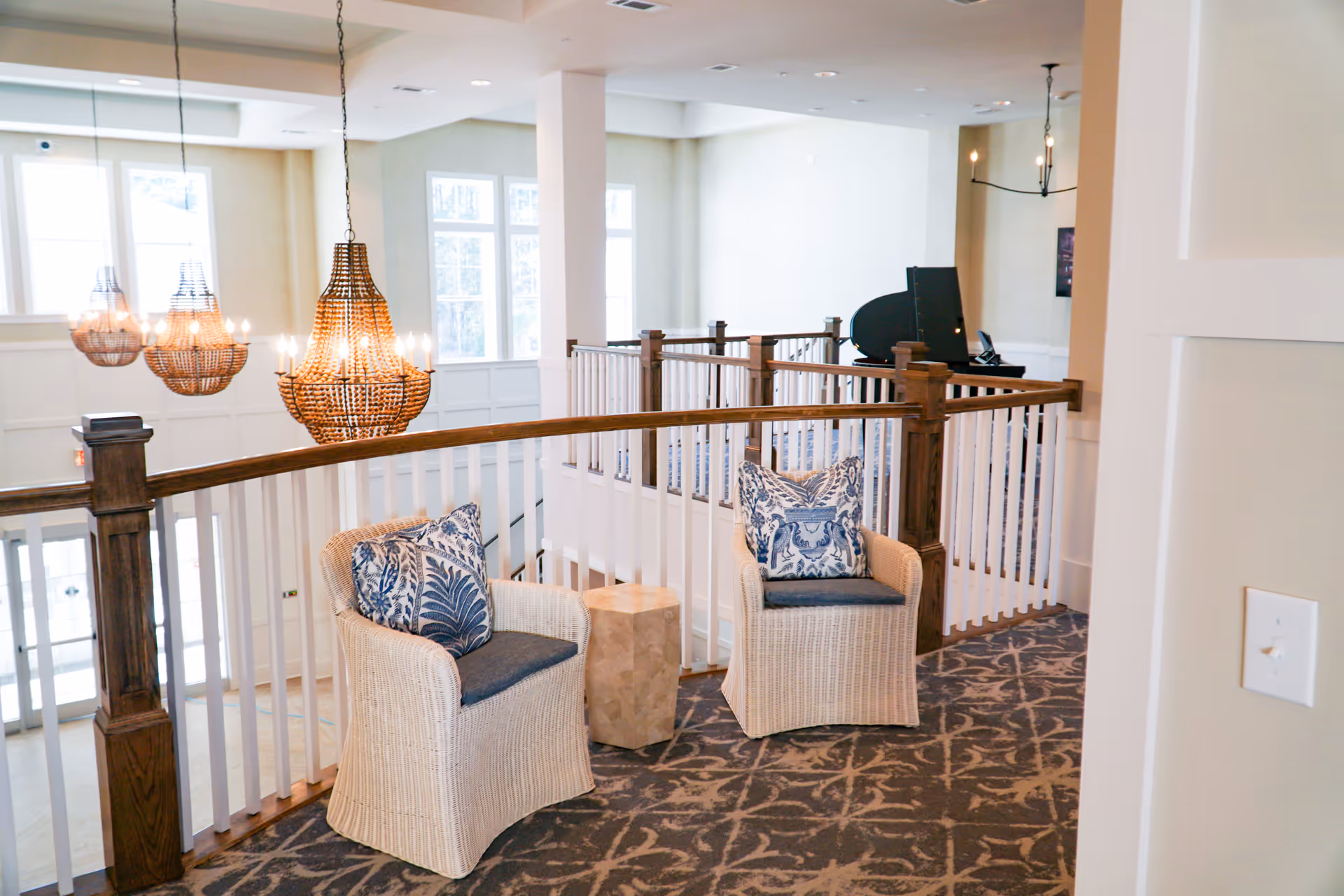 A bright and airy interior seating area with two wicker chairs featuring blue and white patterned cushions, a small wooden side table between them, and a railing overlooking a lower level. Three large, decorative hanging chandeliers are visible, along with large windows letting in natural light and a piano in the background.
