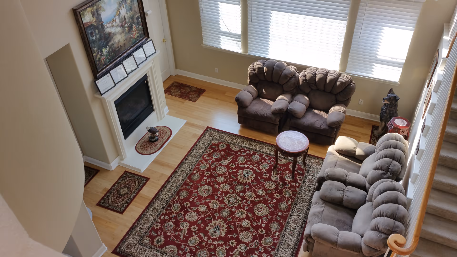 A cozy living room with three plush brown armchairs and a matching sofa arranged around a small round wooden table. The room features a large red patterned area rug on a light wooden floor, a white fireplace with a decorative painting above it, and large windows with white blinds letting in natural light. A staircase with a wooden handrail is visible on the right side.