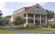 Two-story historic house with a wide columned front porch, American flag, lawn and driveway.