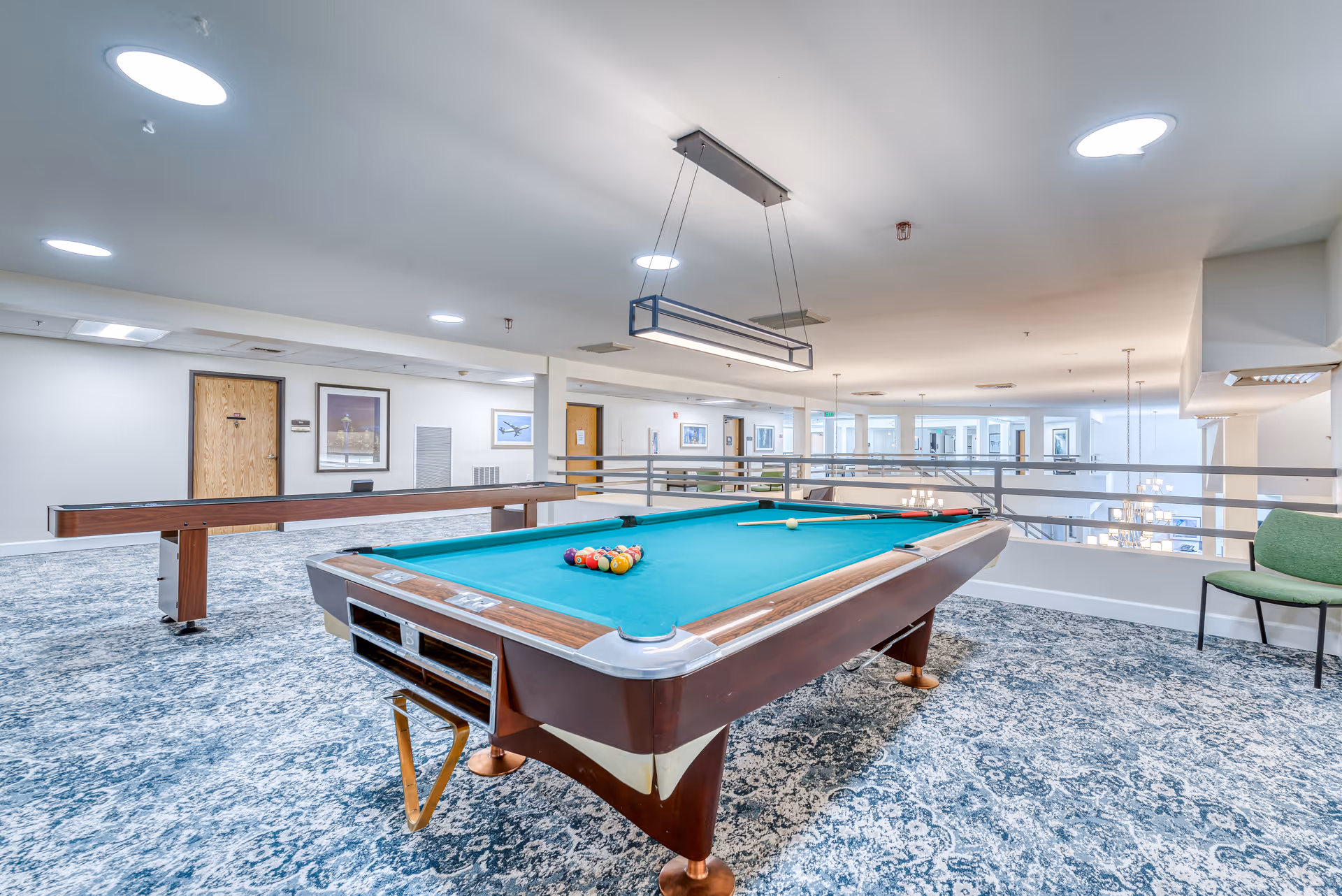 A bright and spacious recreational room featuring a pool table with billiard balls and cues set up for a game. The room has patterned carpet flooring, modern ceiling lights, and a shuffleboard table in the background. There are framed pictures on the walls and a green chair near the railing overlooking a lower level with chandeliers.