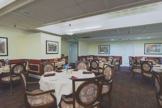 Dining room with multiple round tables covered with white tablecloths, each set with folded napkins, plates, and small flower arrangements. The room has patterned chairs, framed artwork on the walls, and a carpeted floor.