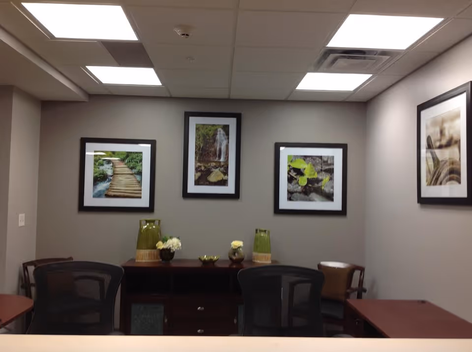 An interior office or reception area with two black mesh chairs and a wooden desk. On the wall behind the desk are four framed nature photographs depicting a wooden bridge over water, a waterfall, green leaves on rocks, and a close-up of a plant. The ceiling has recessed lighting panels.