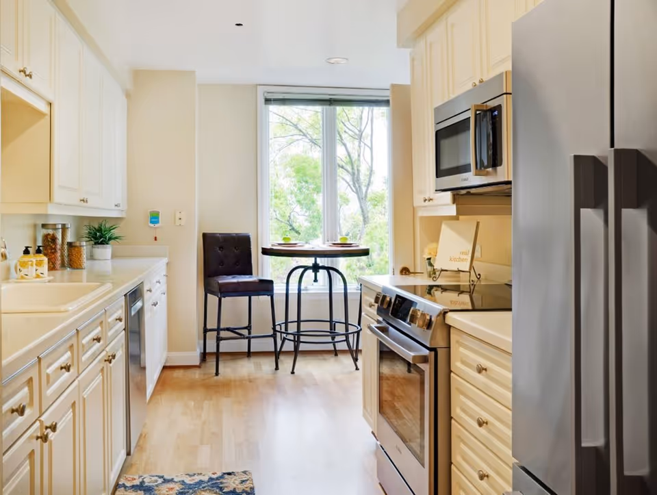 Bright kitchen with white cabinetry, stainless-steel refrigerator and stove, and a small round table with two chairs by a window.