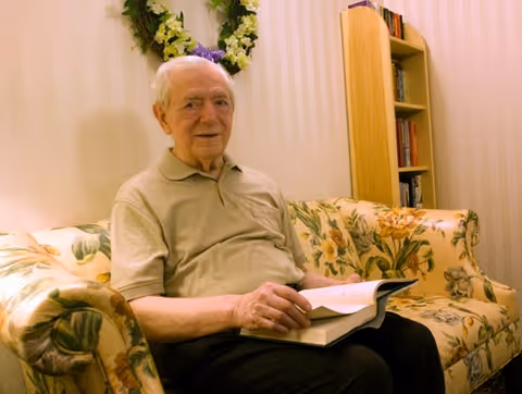An elderly man sitting on a floral-patterned couch in a cozy room, holding an open book. Behind him is a wooden bookshelf filled with books and a decorative wreath hanging on the wall.