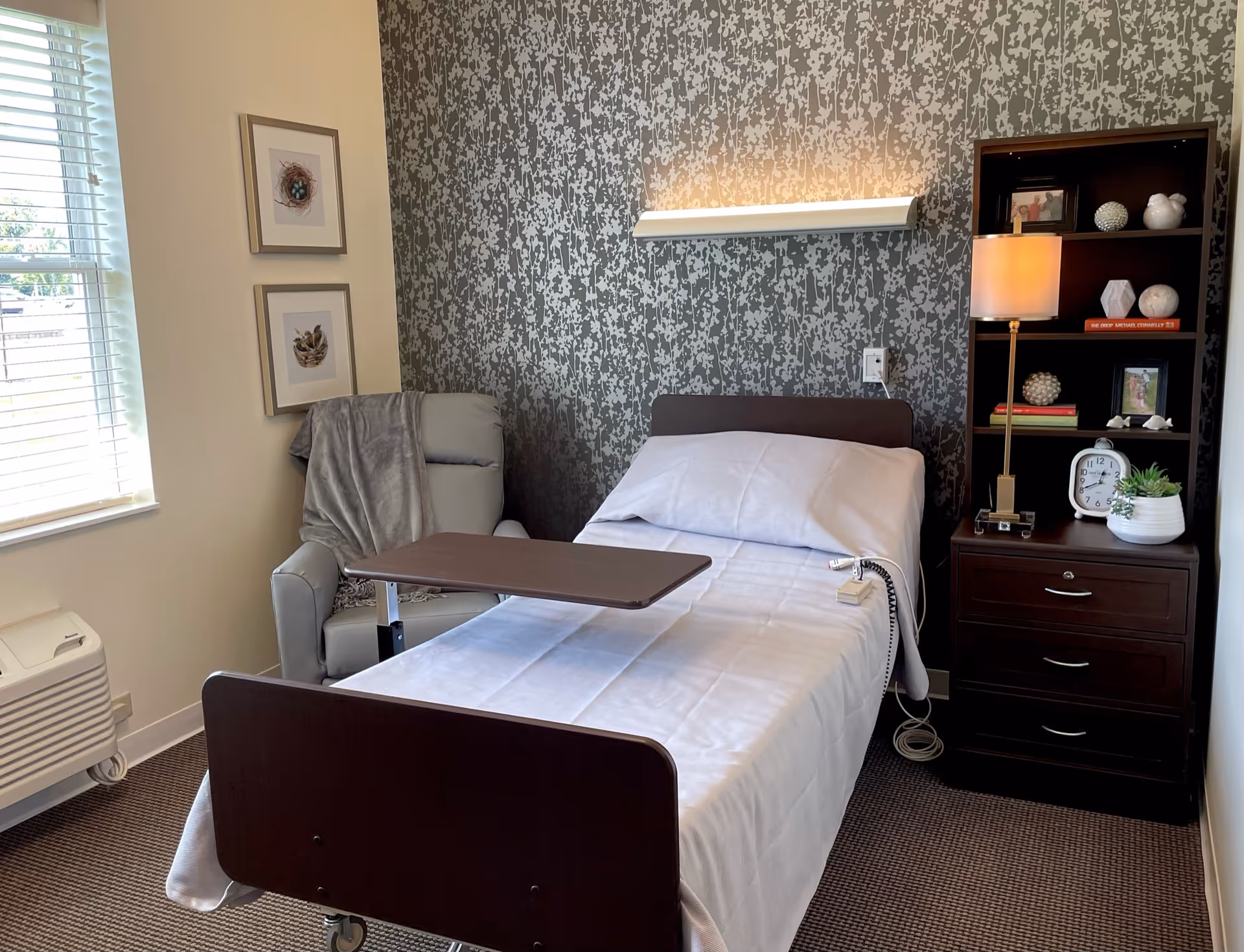 A neatly made hospital-style bed with white linens and an adjustable overbed table in a room. To the left of the bed is a comfortable gray recliner chair with a gray throw blanket draped over it. Behind the bed is a wall with patterned wallpaper and a horizontal light fixture above the headboard. To the right of the bed is a dark wooden nightstand with drawers, a table lamp, a clock, books, framed photos, and decorative items. A window with blinds is on the left side of the room, letting in natural light.