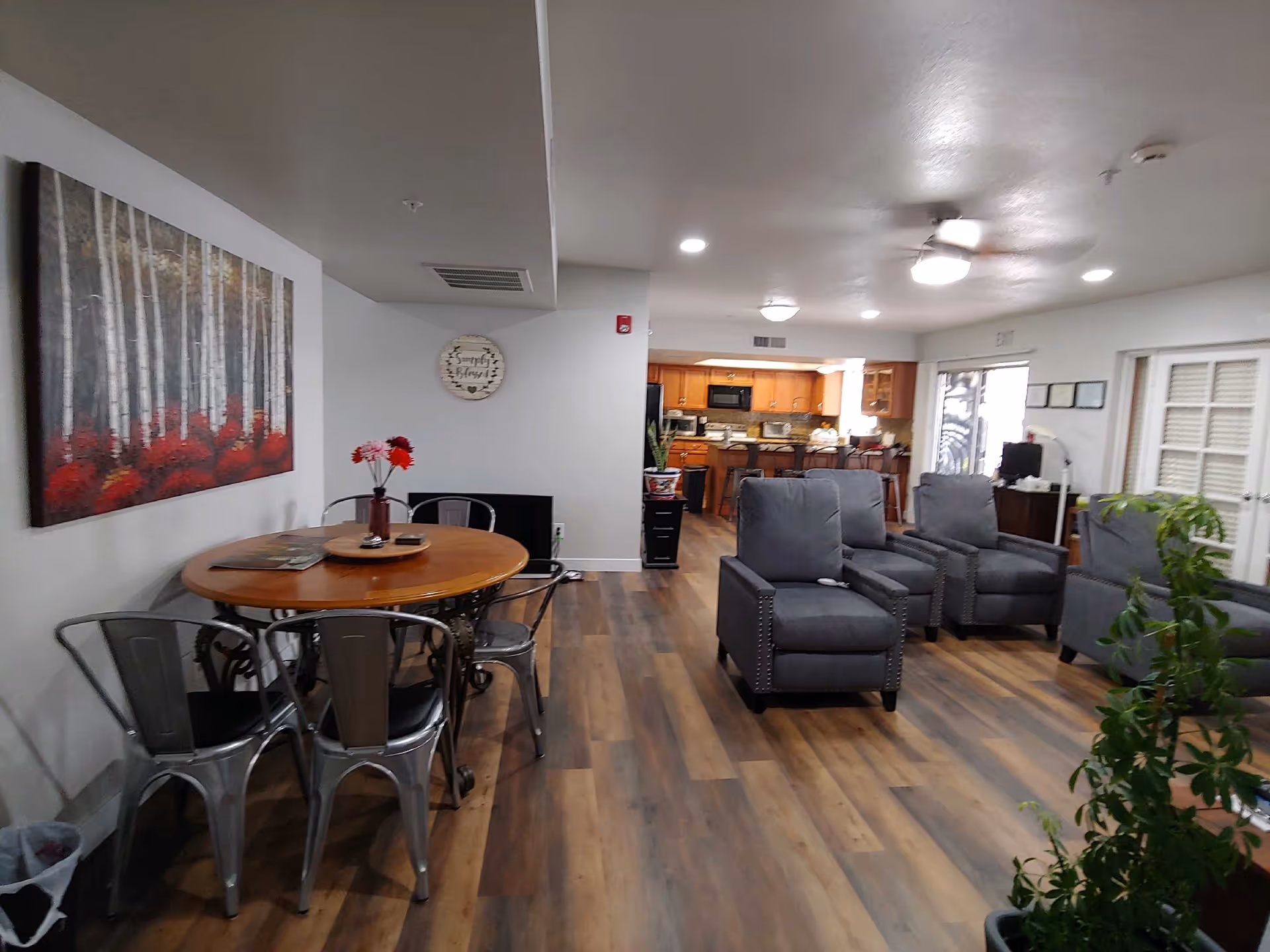 Interior view of a senior living facility common area featuring a round wooden table with metal chairs and a vase with flowers on it, gray upholstered armchairs arranged in a seating area, wooden flooring, a kitchen with wooden cabinets in the background, and wall decorations including a painting of birch trees and a round wall clock.