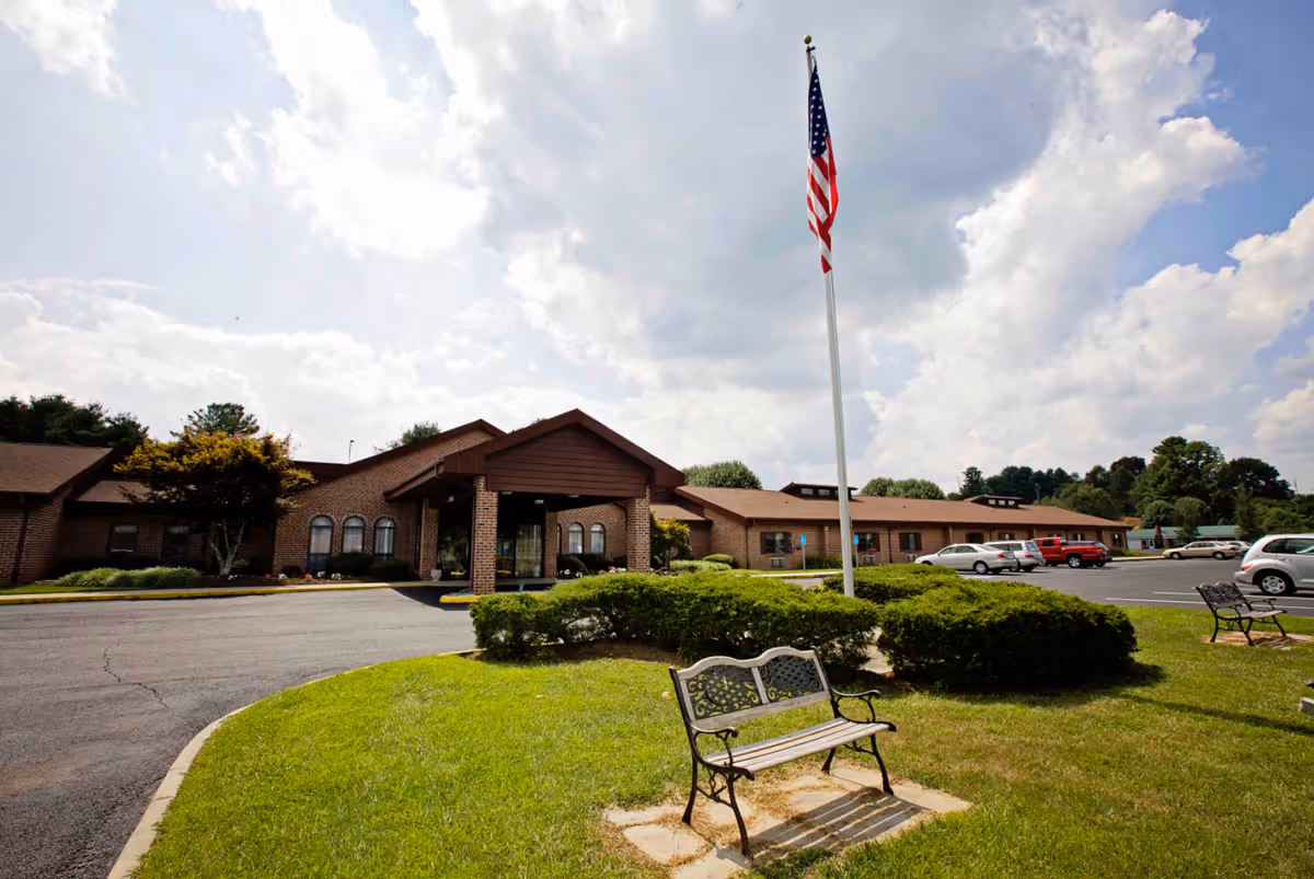 Exterior view of Commonwealth Senior Living At Hillsville, showing a single-story brick building with a covered entrance, an American flag on a tall flagpole, green bushes, a grassy area with benches, and a parking lot with several cars under a partly cloudy sky.