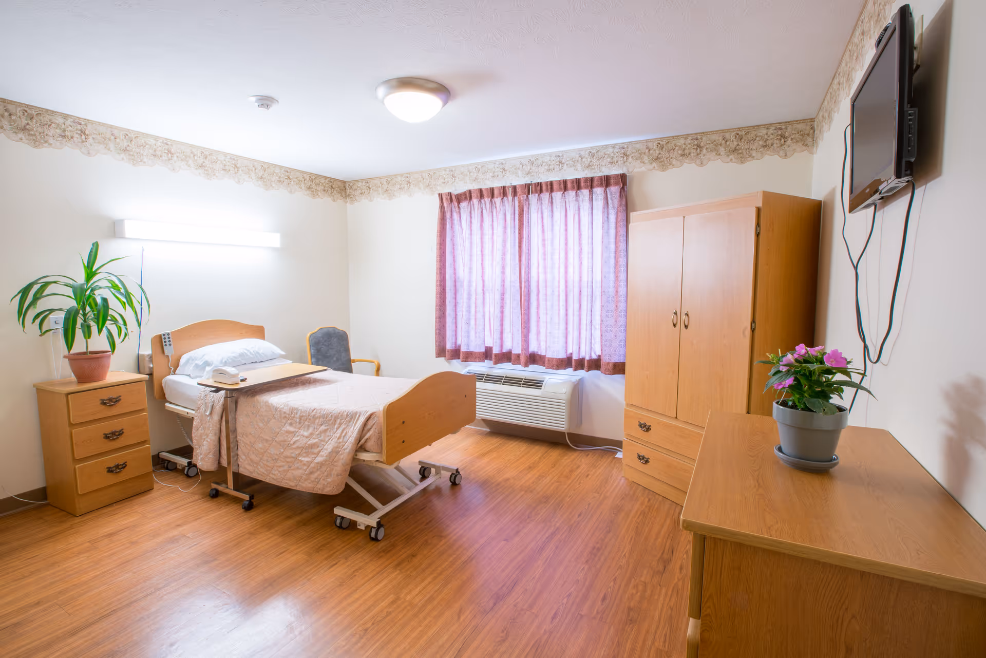 A clean and well-lit bedroom in a senior living facility featuring a single hospital-style bed with a beige quilt, a bedside table with a potted plant, a chair, a wooden wardrobe, a wooden dresser with a potted flower, a wall-mounted TV, and a window with purple curtains. The room has wooden flooring and light-colored walls with decorative border trim near the ceiling.