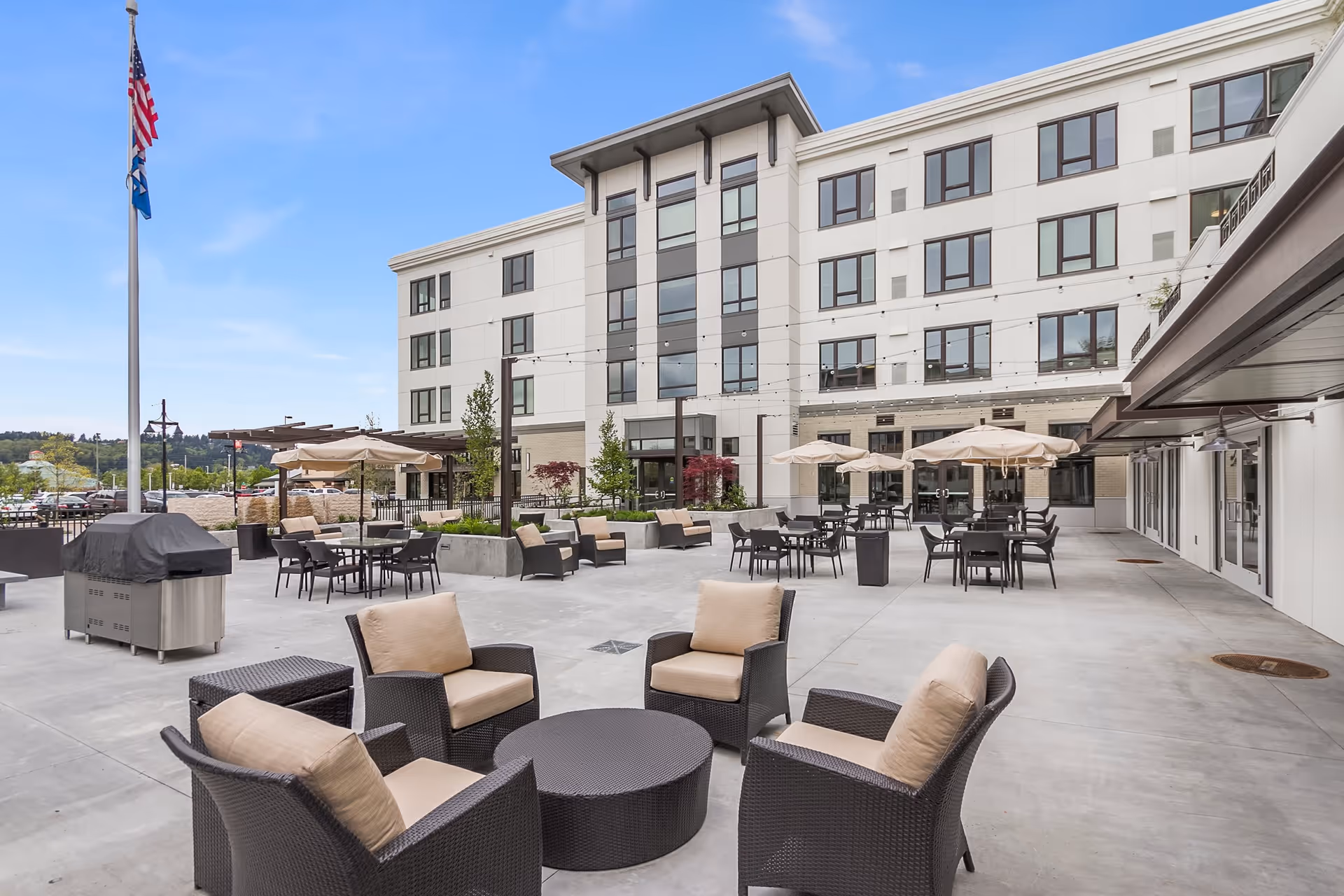 Outdoor patio area at Merrill Gardens at Auburn with multiple seating arrangements including cushioned chairs around a round table, tables with umbrellas, and a flagpole with American and state flags. The background shows a multi-story building with many windows under a clear blue sky.