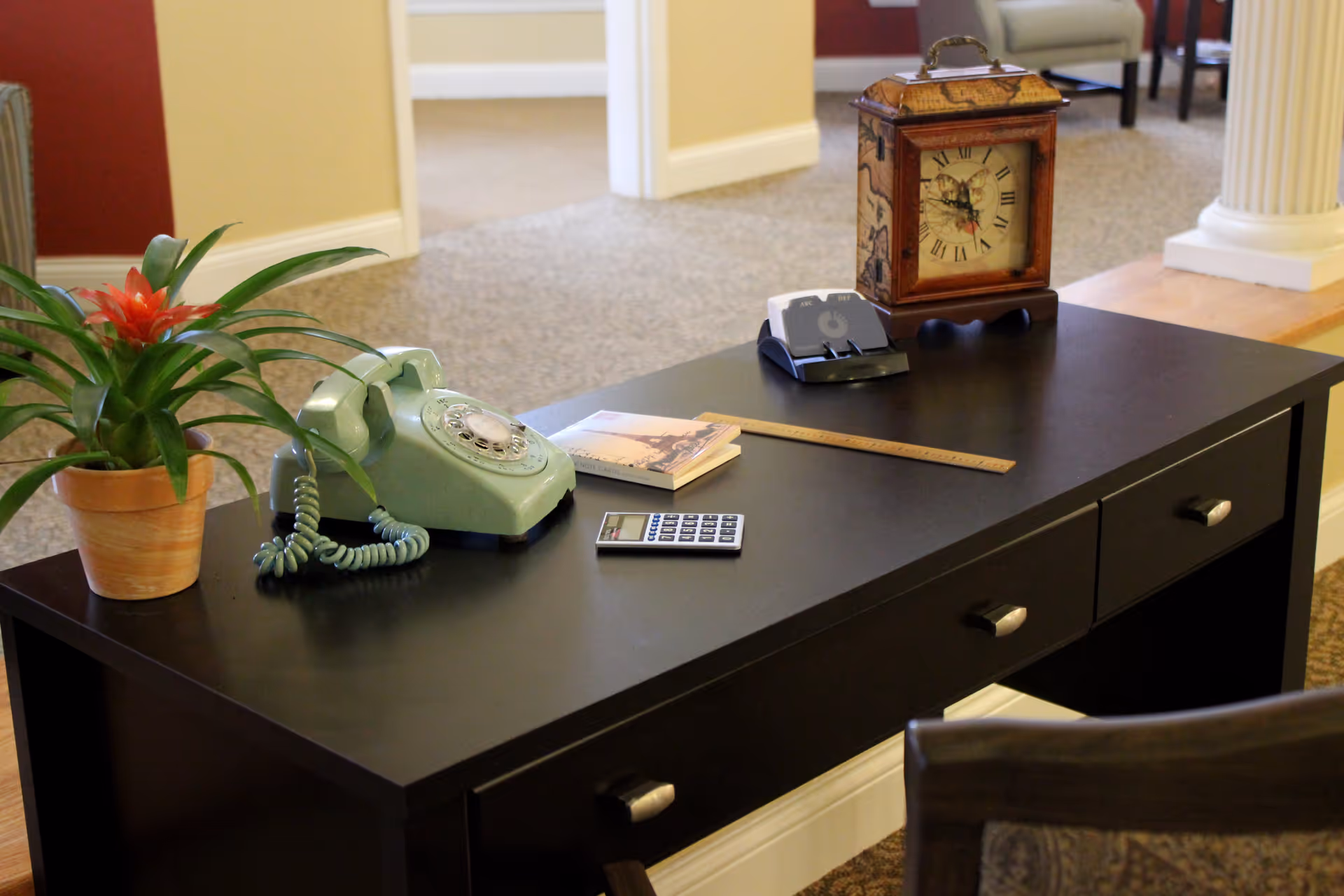 A black desk with various items on it including a potted plant with a red flower, a vintage rotary telephone, a book, a wooden ruler, a calculator, a clock with Roman numerals, and a small electronic device. The background shows a carpeted floor and part of a room with beige walls and white trim.