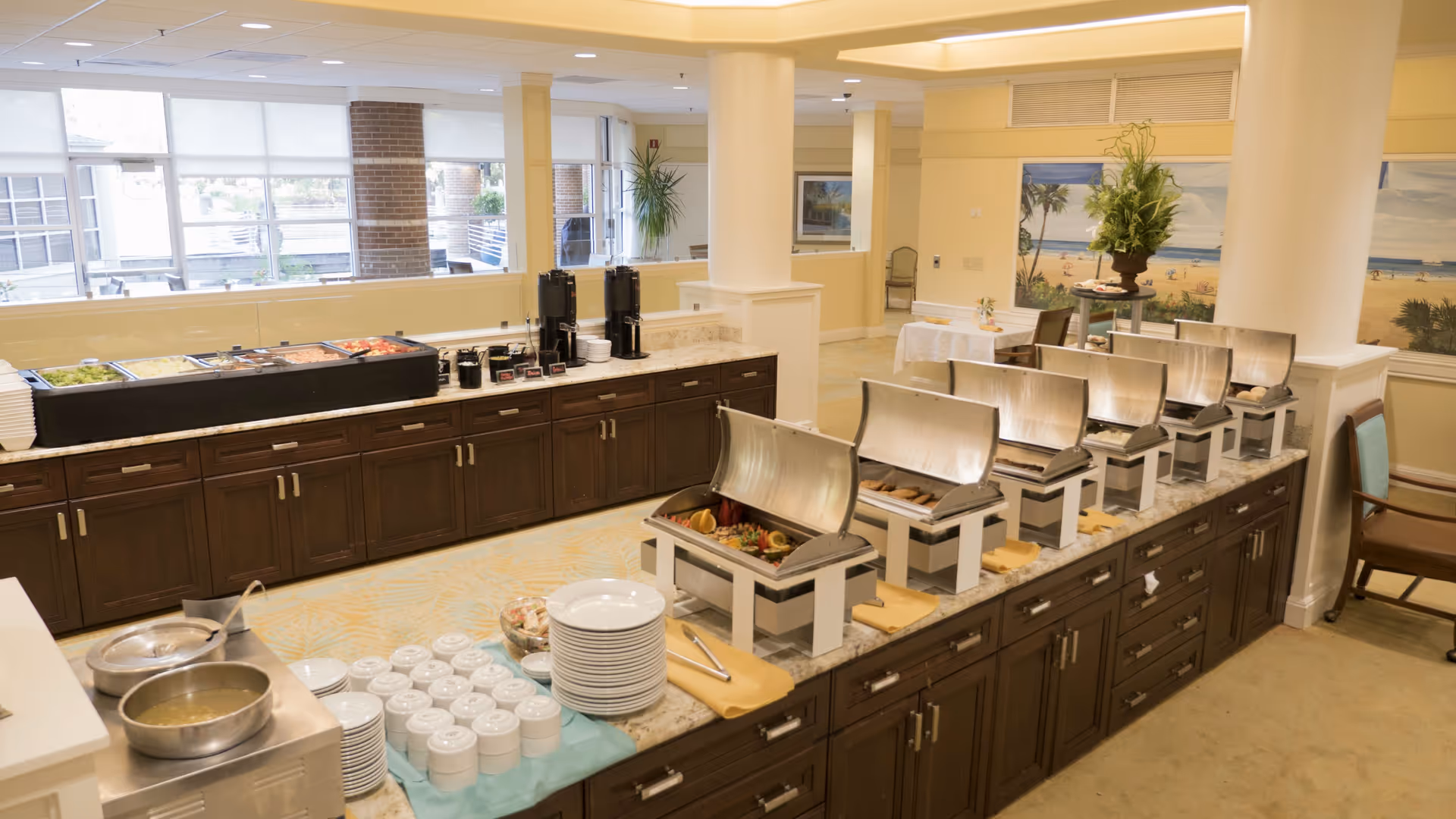 Buffet-style dining area with multiple chafing dishes containing food, stacks of plates and bowls, coffee dispensers, and a salad bar in a well-lit room with large windows and yellow walls.