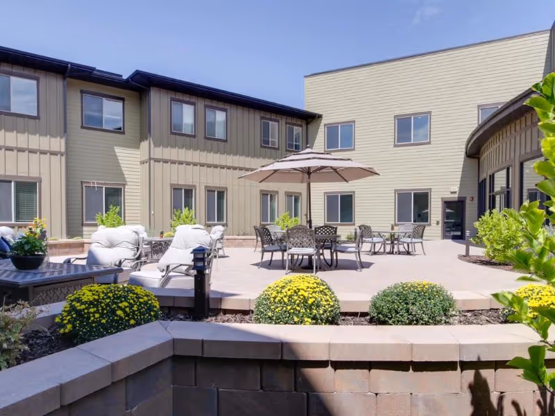 Outdoor patio area at Spring Gardens Senior Living Draper with cushioned chairs, tables with umbrellas, and decorative plants surrounded by a two-story building.