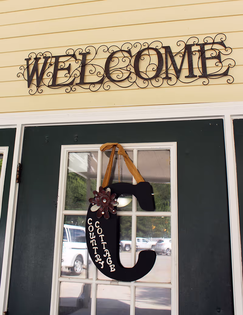 A dark green door with glass panes has a large decorative letter 'C' hanging on it, adorned with a flower and the words 'Country Cottage' written vertically. Above the door, there is a metal wall decoration spelling out 'WELCOME' in ornate letters. The exterior wall is painted yellow, and cars are visible parked outside through the glass panes.