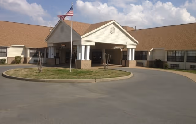 Front exterior view of a single-story healthcare facility building with a covered entrance supported by white columns, an American flag on a flagpole in front, and a circular driveway.