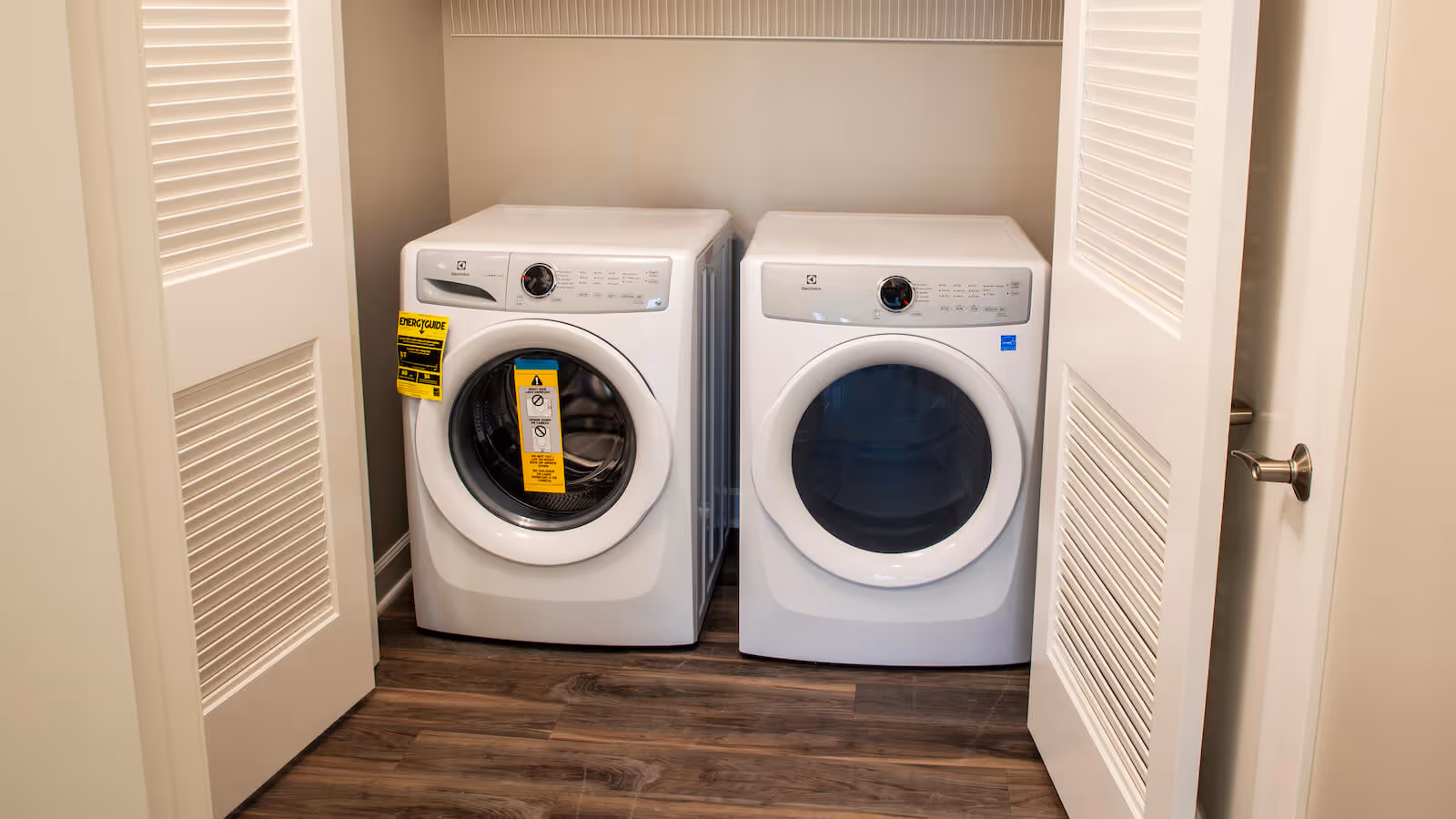 A laundry area with a white front-loading washing machine and a matching dryer side by side, enclosed by white louvered closet doors, with wood flooring underneath.