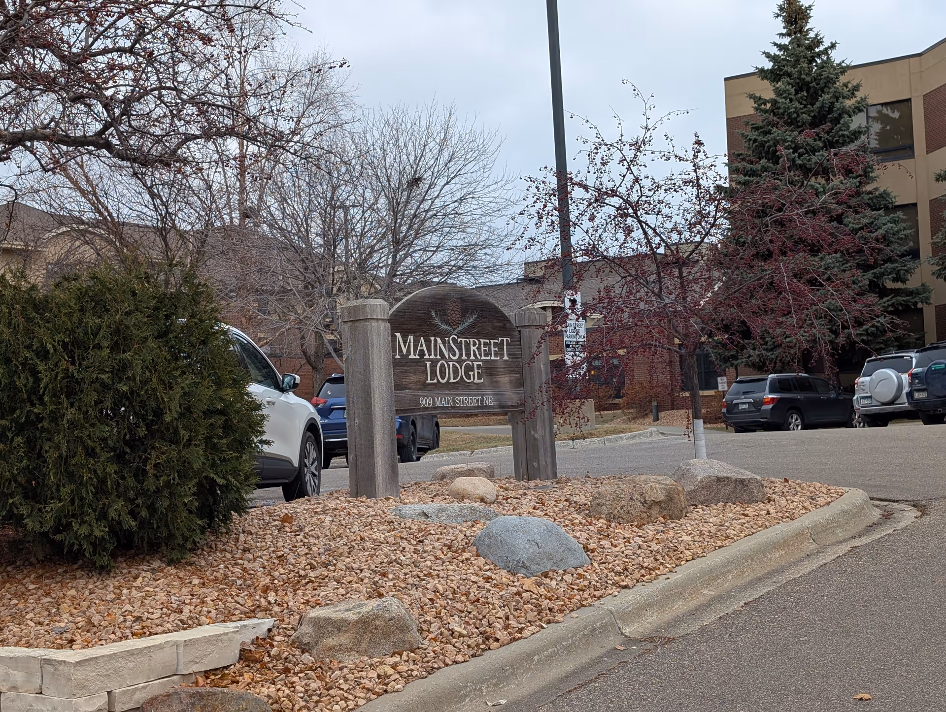 Wooden 'MainStreet Lodge' sign in a rock-mulched island at the entrance of a parking lot with the lodge building and parked cars in the background.