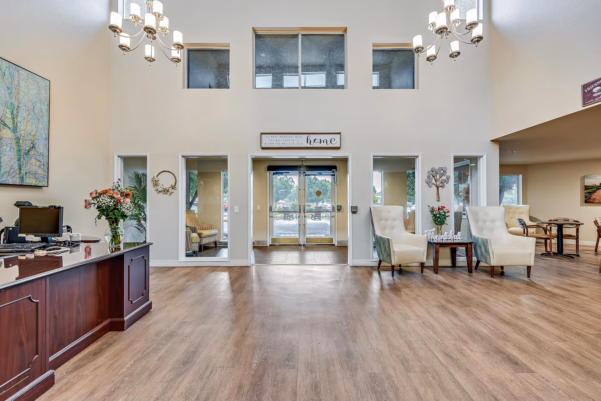 Spacious senior living facility lobby with high ceilings, wooden flooring, and large glass entrance doors. The area features a reception desk with a computer and flowers on the left, two cream-colored armchairs with a small table and flowers between them on the right, and several windows allowing natural light. Decorative wall art and chandeliers hang from the ceiling.