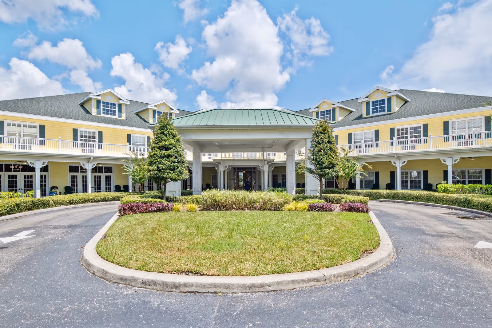 Front exterior view of a two-story senior living facility building with yellow siding, white trim, and a green roof. There is a circular driveway with landscaped greenery and trees in the center, under a partly cloudy blue sky.