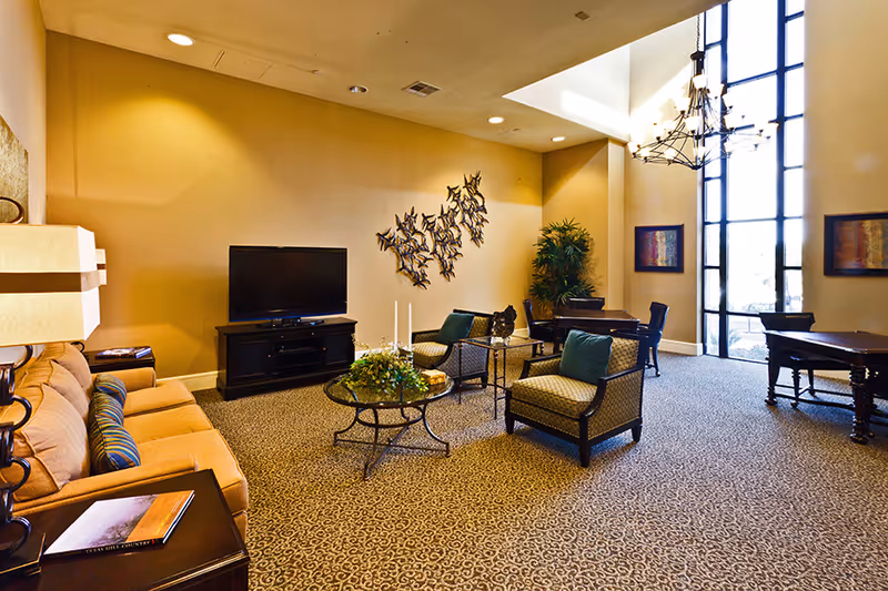 A spacious living room area in an assisted living facility with a beige sofa, two armchairs with green cushions, a glass coffee table with decorative items, a flat-screen TV on a black stand, and large windows letting in natural light. The room features patterned carpet, wall art, a chandelier, and additional tables and chairs near the windows.