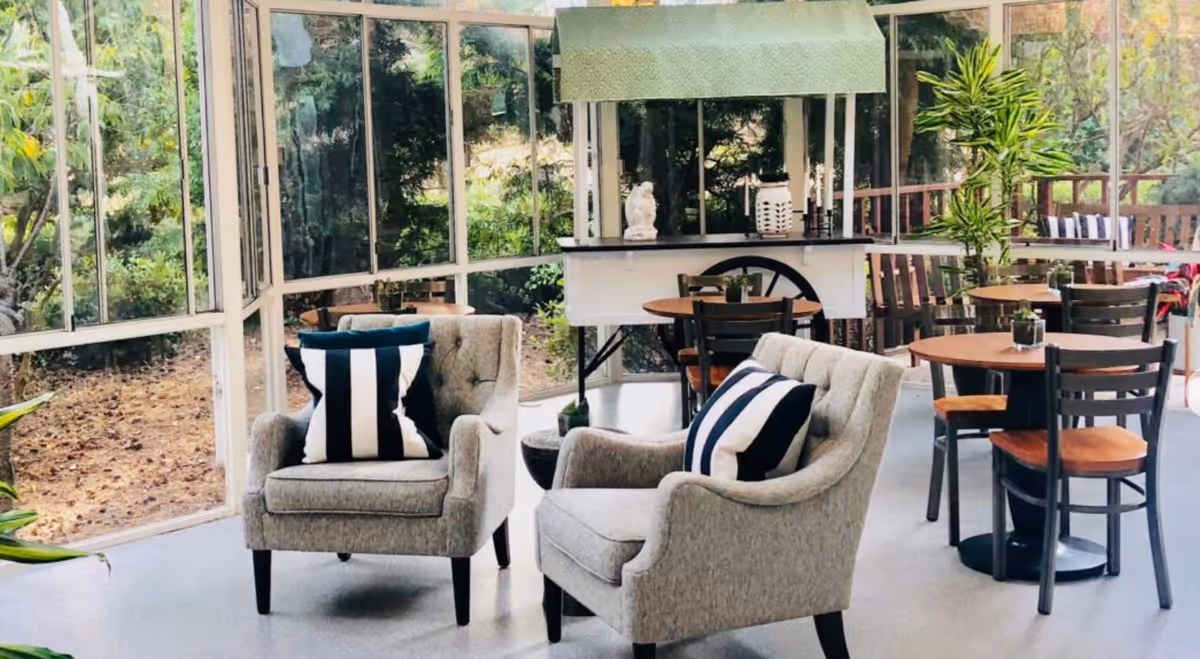 A bright and airy seating area with two gray upholstered armchairs each with a black and white striped pillow, several round wooden tables with black chairs, large windows showing greenery outside, and a white console table with decorative items.
