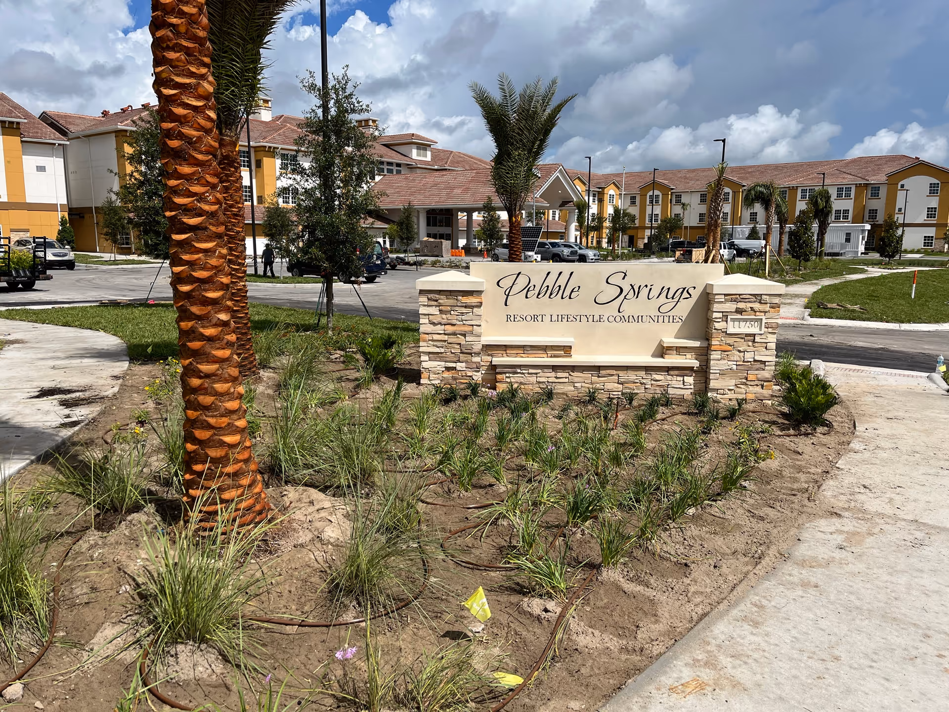 Entrance sign for Pebble Springs Resort Lifestyle Communities surrounded by newly planted landscaping and palm trees, with a large building in the background under a partly cloudy sky.