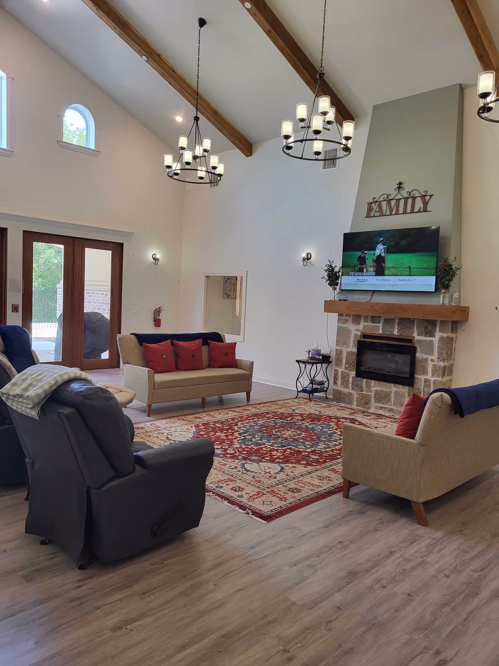 A spacious living room with high vaulted ceilings featuring wooden beams and two circular chandeliers. The room has a stone fireplace with a mounted TV above it displaying a scene from a movie. Above the TV, there is a decorative sign that reads 'FAMILY'. The seating includes two beige sofas with red cushions and two dark recliner chairs. A large patterned area rug covers part of the wooden floor. There are large glass doors and small arched windows letting in natural light.