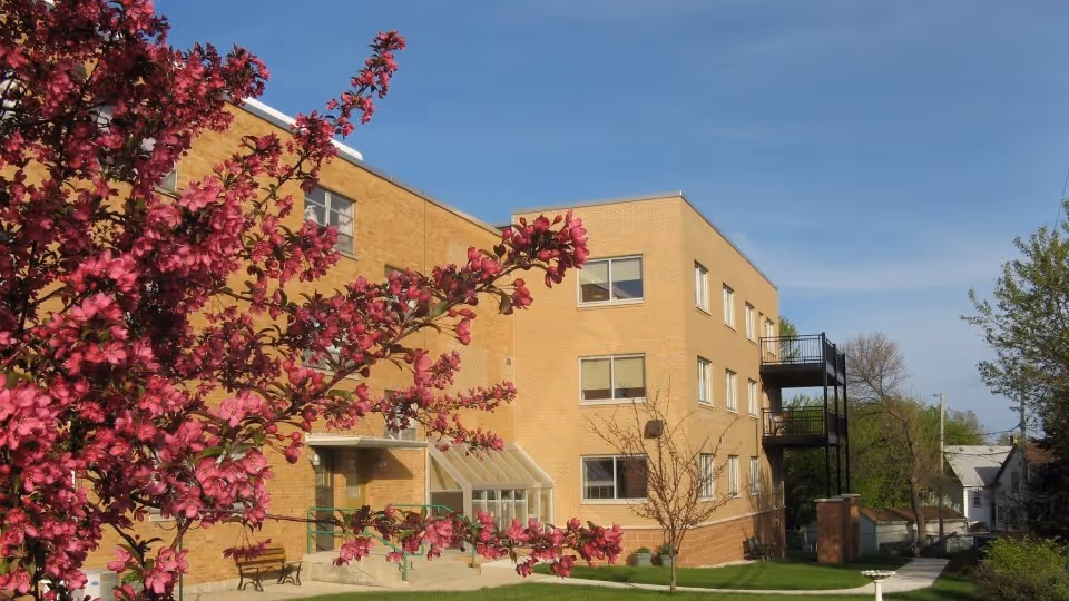 Exterior view of a three-story brick building with multiple windows, a small entrance with a glass awning, and an external metal staircase. In the foreground, there is a tree with vibrant pink blossoms and a well-maintained lawn with a few small trees. The sky is clear and blue.