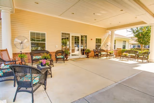 Covered outdoor patio area with wicker chairs and tables arranged for seating. There are two large fans mounted on the wall, potted plants, and a double glass door leading inside the building. The area is bright and sunny with trees visible in the background.