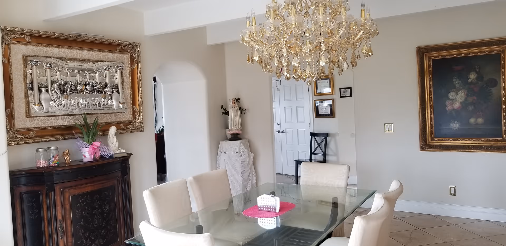 Bright dining room with a glass table surrounded by upholstered chairs, a crystal chandelier, ornate framed artwork, and a wooden cabinet.