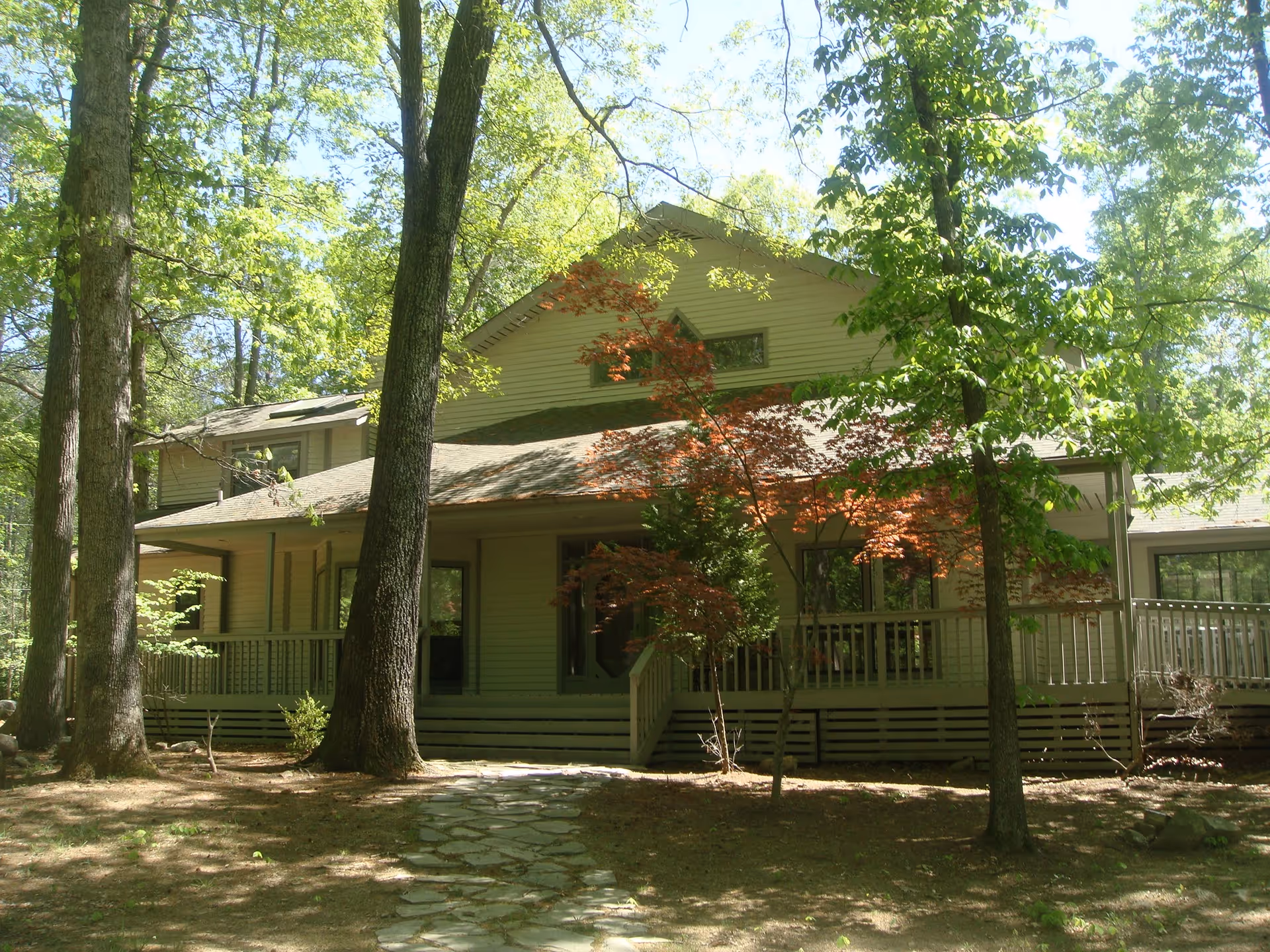 A two-story house surrounded by tall trees and greenery with a stone pathway leading to the front porch. The house has a light-colored exterior and a sloped roof, with a wooden railing around the porch.