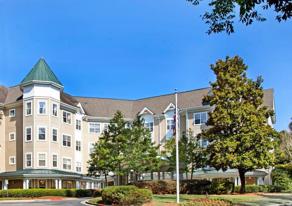 Front view of a multi-story senior living building with a turret, flagpole, and landscaped trees under a clear blue sky.